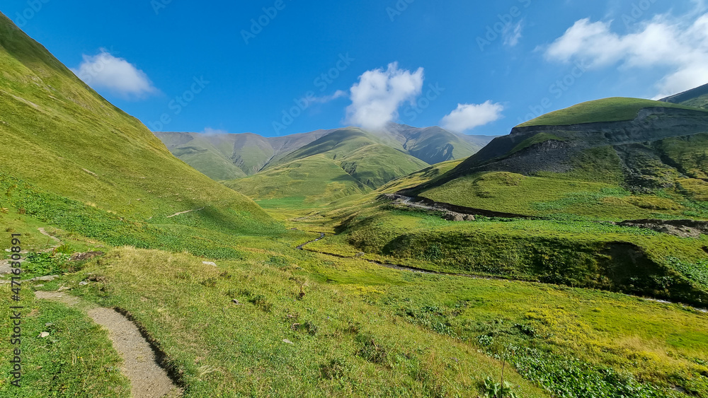 A hiking trail leading through a vast green grassland in the Greater Caucasus Mountain Range in Georgia, Kazbegi Region. The surrounding mountains and hills are green and soft. Tranquility. Highlands