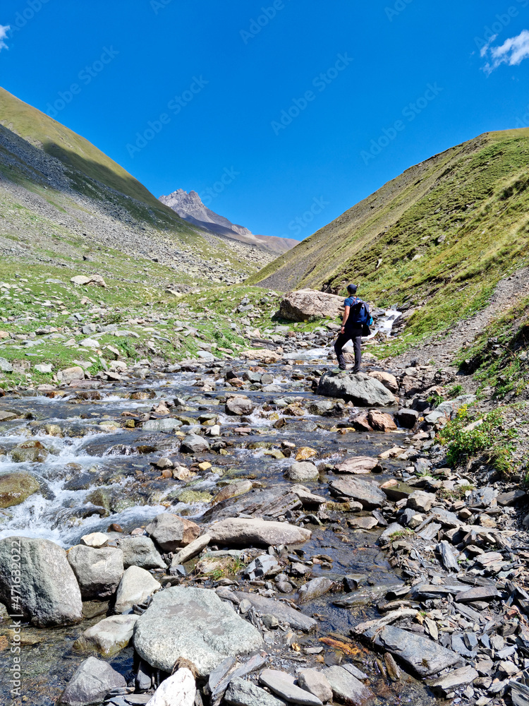 A man walking on a hiking trail next to a river with a view on the sharp mountain peaks of the Chaukhi massif in the Greater Caucasus Mountain Range in Georgia, Kazbegi Region. Remedy, Wanderlust.