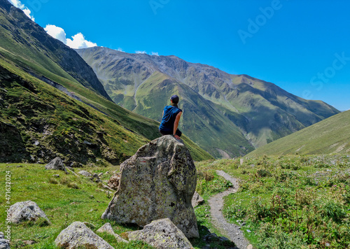 A male hiker sitting on a massive rock next to a hiking trail and enjoying the panoramic view on the high ridges of the mountain peaks in the Greater Caucasus Mountain Range in Georgia,Kazbegi Region.