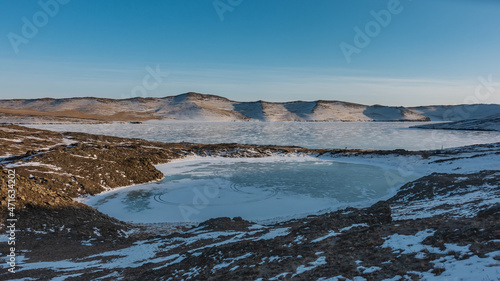 A small frozen lake in the shape of a heart is separated from the big one by a strip of land. Snow on the banks. A mountain range against a blue sky. Baikal.