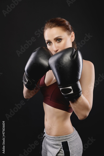 Caucasian woman boxer in sportswear and boxing gloves stands in a fighting stance and looks into the camera. Against a dark background. Strong woman's female fights