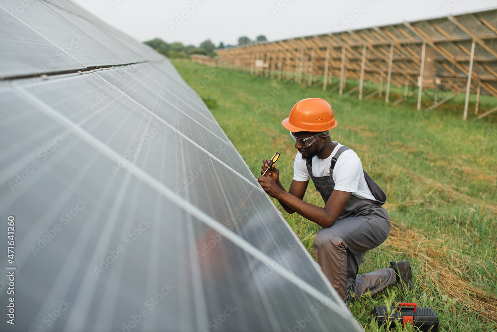 African american man in uniform and helmet using screwdriver for repairing solar panels on station. Installation of photovoltaic cells. Green energy concept. African american man in uniform and helmet using screwdriver for repairing solar panels on station. Installation of photovoltaic cells. Green energy concept.
