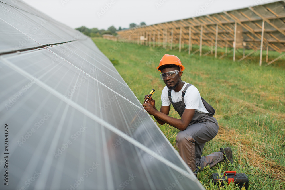 African american man in uniform and helmet using screwdriver for repairing solar panels on station. Installation of photovoltaic cells. Green energy concept. African american man in uniform and helmet using screwdriver for repairing solar panels on station. Installation of photovoltaic cells. Green energy concept.