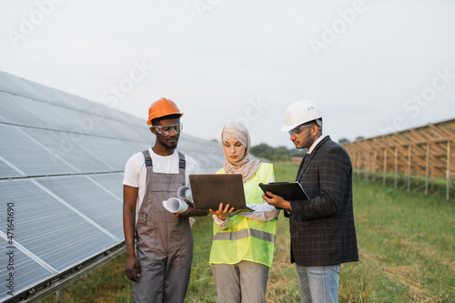 Muslim woman in hijab and indian man in white helmet talking with african american technician on solar station. Multiracial people using laptop, clipboard and blueprints for work outdoors.
