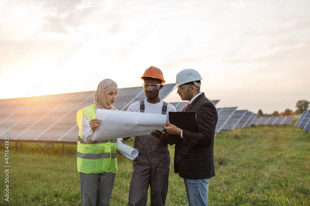 Group of three multiracial people in safety helmets having working meeting among solar station. Technician with engineers standing outdoors with blueprints in hands. Group of three multiracial people in safety helmets having working meeting among solar station. Technician with engineers standing outdoors with blueprints in hands.