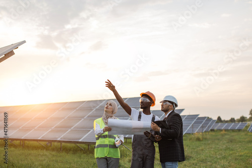 African american technician talking with muslim woman and indian man during working session on solar station. Industrial workers discussing common project of green energy production.