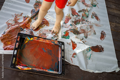 Little boy draws with hands and feet with finger paints on a large sheet at home