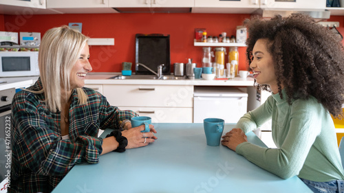 Interracial young girls talking sitting on the young hostel common space holding cups of coffee and milk