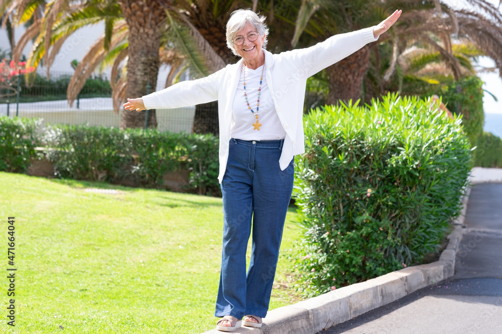 Happy senior woman walks on the curb with her arms outstretched to keep balance. Playful elderly retiree outdoor in a garden