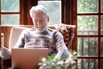 Smiling senior man in winter sweater sitting in living room using laptop computer. Carefree elderly grandfather enjoying tech and social. Rustic chalet in the wood
