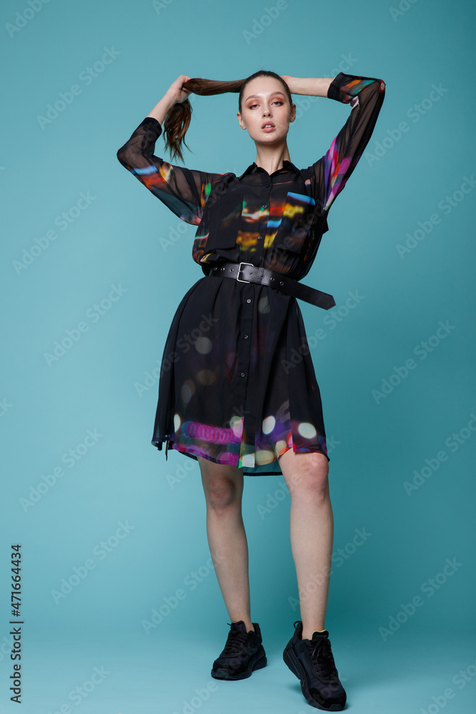 High fashion photo of a beautiful elegant young woman in a pretty black dress with colored patterns, sneakers posing over turquoise, blue background. Studio Shot. 
