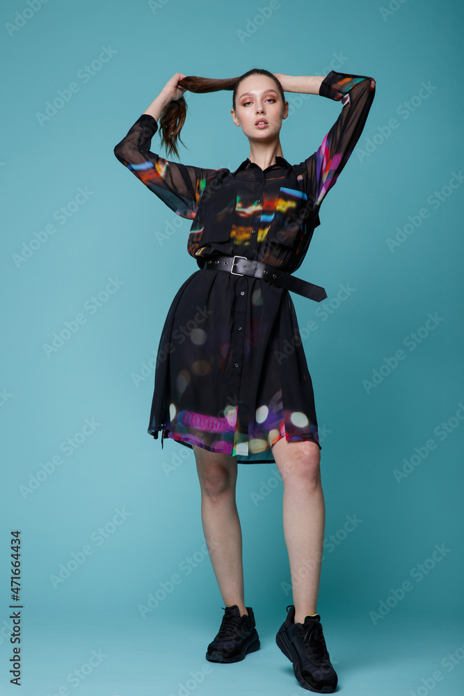 High fashion photo of a beautiful elegant young woman in a pretty black dress with colored patterns, sneakers posing over turquoise, blue background. Studio Shot. High fashion photo of a beautiful elegant young woman in a pretty black dress with colored patterns, sneakers posing over turquoise, blue background. Studio Shot.