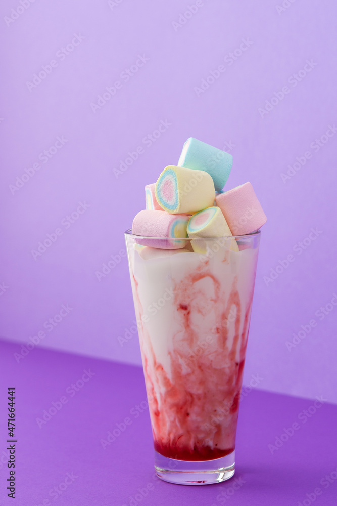 Glass cup with milkshake and fruit syrup, multicolored marshmallows on bright purple background, holiday or birthday concept Glass cup with milkshake and fruit syrup, multicolored marshmallows on bright purple background, holiday or birthday concept