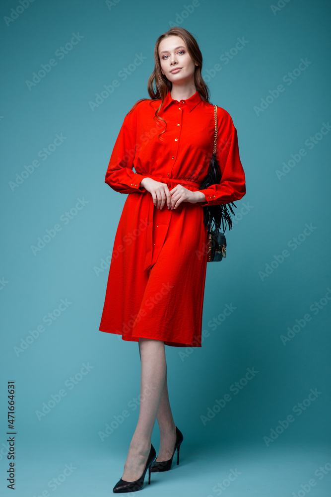 High fashion photo of a beautiful elegant young woman in a pretty red dress, black handbag posing over turquoise, blue background. Studio Shot.