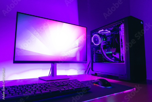 Modern gaming computer setup with display, water cooling case, keyboard, mouse on gaming mat. Purple led light in background