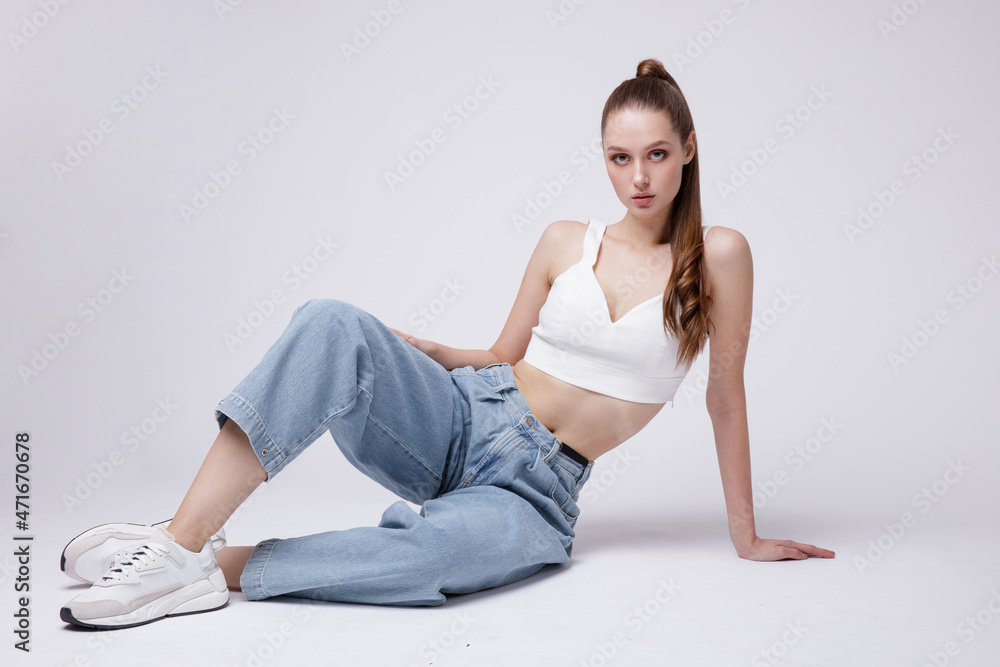High fashion photo of a beautiful elegant young woman in a pretty white top and sneakers, blue denim jeans posing over white, soft gray background. Studio Shot. The model is sitting High fashion photo of a beautiful elegant young woman in a pretty white top and sneakers, blue denim jeans posing over white, soft gray background. Studio Shot. The model is sitting