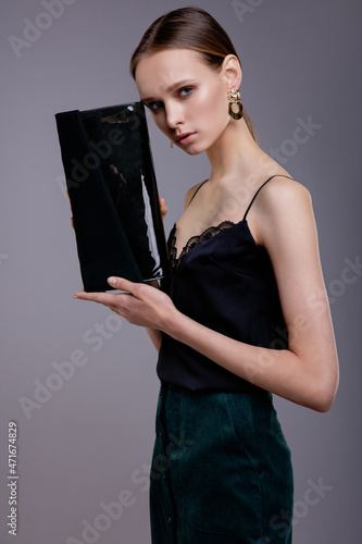 Fashion model in black top and green (emerald scarab) skirt, with a clutch in hand, beautiful young woman. Studio shot. Gray background. 