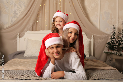 a young family with a little daughter in Santa Claus hats are lying on the bed at home Christmas