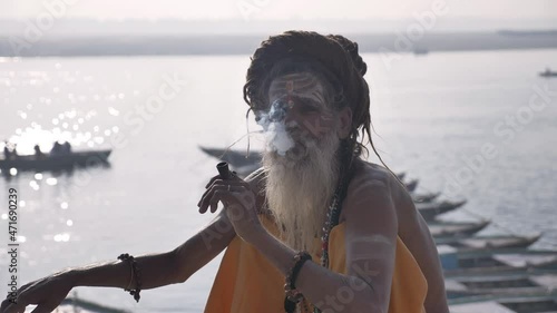 A bearded old or elderly sadhu sitting and smoking on the banks of the Ganga in Varanasi, India.  photo