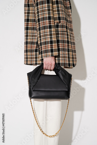 Woman in plaid blazer and white leather pants with black leather handbag with golden chain in hand. White background. Vertical photo.