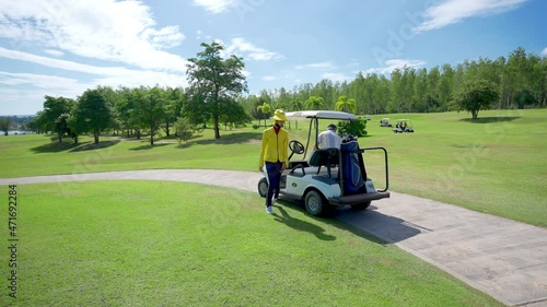 4K Asian senior man with female caddy driving golf cart together on golf course to fairway at country club. Healthy elderly male golfer walking to golfing on the green in summer sunny day photo