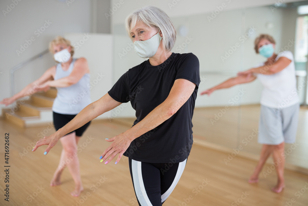 Positive mature woman wearing protective face mask enjoying active dancing during group training in choreographic studio. Precautions in coronavirus pandemic