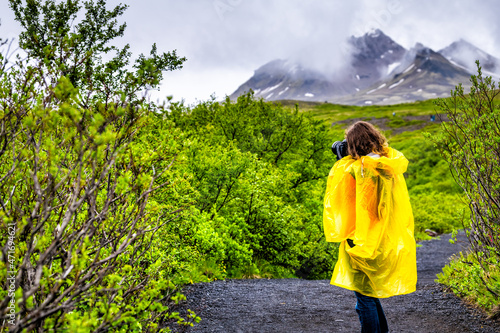 Skaftafell, Iceland green summer lush landscape view of woman tourist in yellow poncho walking hiking on wet trail path hiking road rainy day in mountains