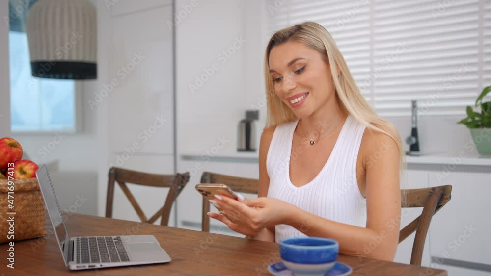 Young beautiful woman sits at the table in her kitchen drinking coffee and texting with her friends Young beautiful woman sits at the table in her kitchen drinking coffee and texting with her friends