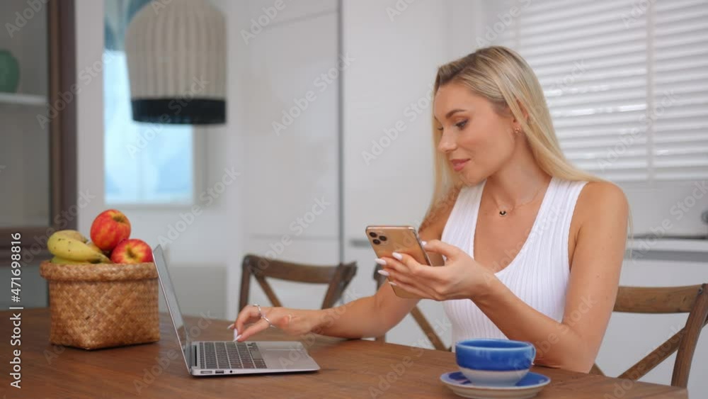 Pretty woman in white top with phone in hand sits at the table working from home, typing in laptop Pretty woman in white top with phone in hand sits at the table working from home, typing in laptop