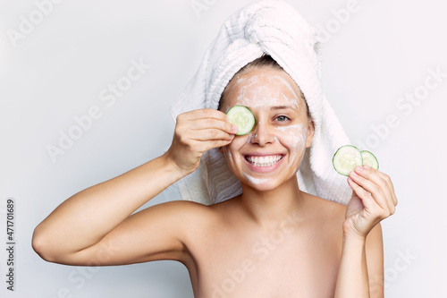 A young happy smiling woman with a white towel on her head after a shower holds cucumber slices making a refreshing face mask isolated on a white background. Skin care, cosmetology