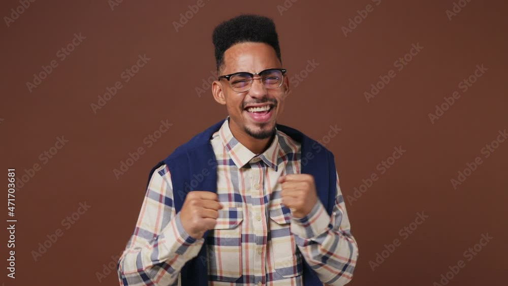 Excited jubilant young african american man 20s wears striped shirt glasses looks around doing winner gesture celebrate clenching fists say yes isolated on plain dark brown background studio portrait Excited jubilant young african american man 20s wears striped shirt glasses looks around doing winner gesture celebrate clenching fists say yes isolated on plain dark brown background studio portrait