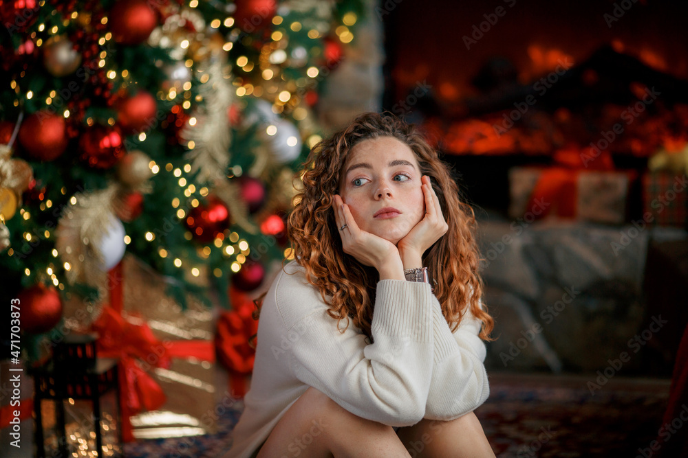 A beautiful girl is sad in the Christmas interior. Christmas tree, lots of gifts, Christmas theme. Cozy atmosphere. Portrait of a red-haired girl on a beautiful background.