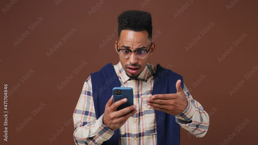 Sad irritated young african american man 20s wear shirt glasses using mobile cell phone swear hears fake news unexpected rumor has some problems isolated on plain dark brown background studio portrait Sad irritated young african american man 20s wear shirt glasses using mobile cell phone swear hears fake news unexpected rumor has some problems isolated on plain dark brown background studio portrait