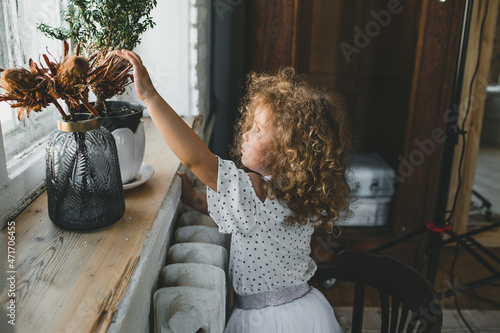 little girl with curly hair touching dried flowers on the windowsill