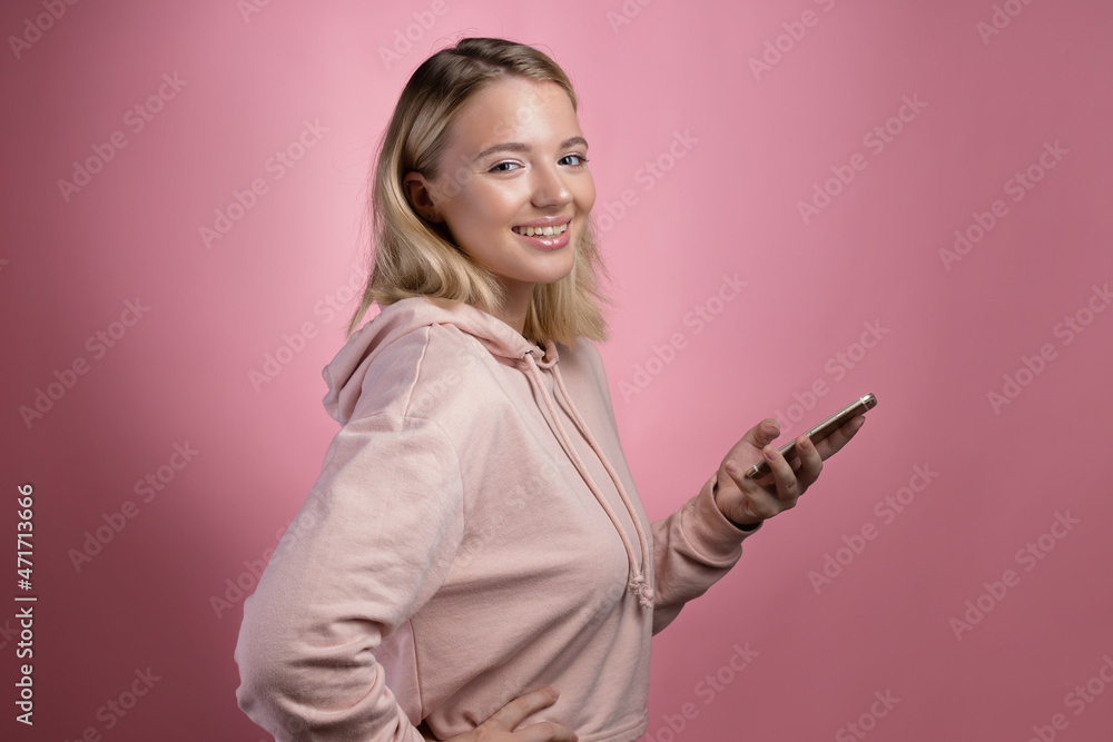 Modern online communication in messengers, correspondence applications. An attractive young woman in a pink hoodie uses a smartphone. A cute blonde holds a phone and looking at you Modern online communication in messengers, correspondence applications. An attractive young woman in a pink hoodie uses a smartphone. A cute blonde holds a phone and looking at you