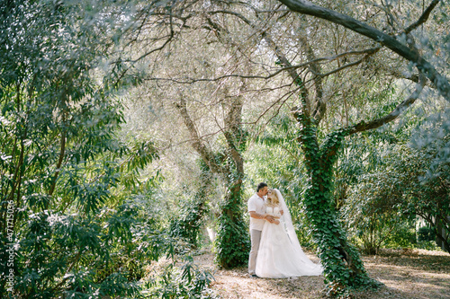 Groom and bride in a white dress stand in the park among the trees