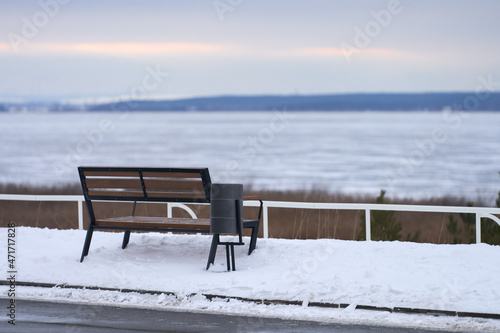 A bench in front of the river on a snowy embankment. The river is covered with thin ice. Selective focus. Copy space.