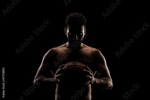 Basketball player holding a ball against black background. Side lit muscular Caucasian man silhouette © Nikola Spasenoski