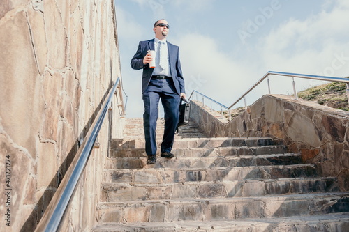 Confident middle age businessman with briefcase walking upstairs. Close-up of businessman wearing blue suit holding bag and coffee cup walking down the stairs