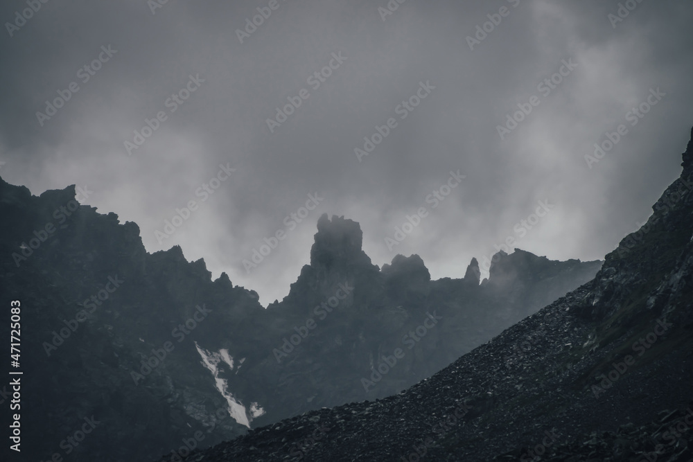 Dark atmospheric mountain landscape with black pointy rocky peak in gray cloudy sky. Lead gray low clouds among black mountains. Dark rocky pinnacle in low clouds in rainy weather. Gloomy minimalism.