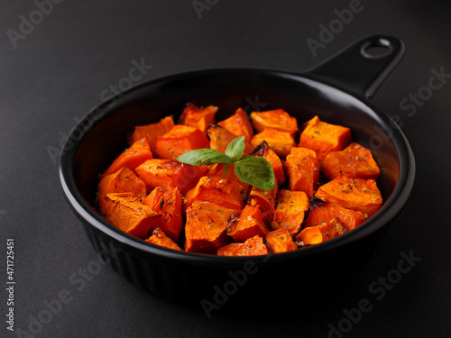 Pumpkin cut into pieces baked in a black round frying pan with a handle on a black background.