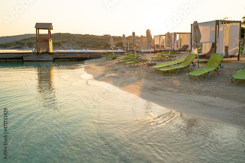 Sandy beach with turquoise sea water, green sun loungers, brown sun beds and a wooden surveillance tower on the jetty in the morning while the sun rises photo