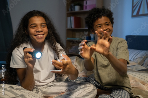 Happy playful African American kids in pijamas sitting on bed in dark room and shining flashlight on wall while making hand shadow of creatures