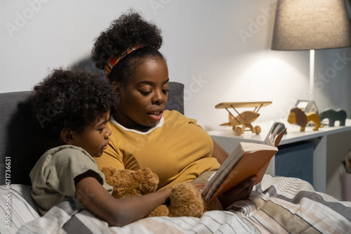 African American mother with curly hair sitting in bed and reading book to son while spending time with him before bedtime