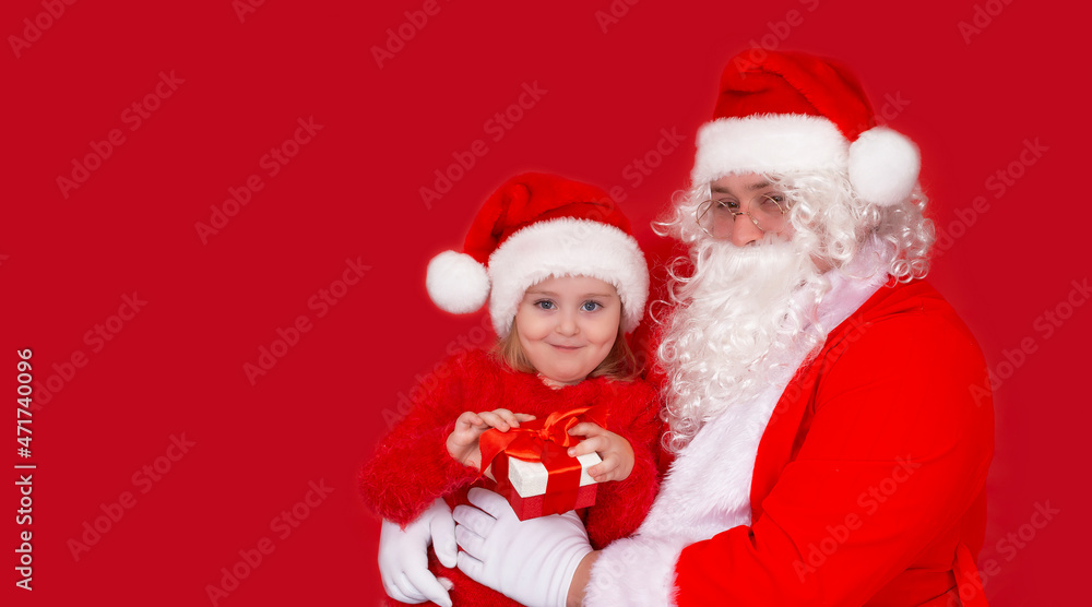 Smiling little girl in a hat with Santa Claus and gifts, isolate on a red background. Holidays, childhood and the concept of people Smiling little girl in a hat with Santa Claus and gifts, isolate on a red background. Holidays, childhood and the concept of people