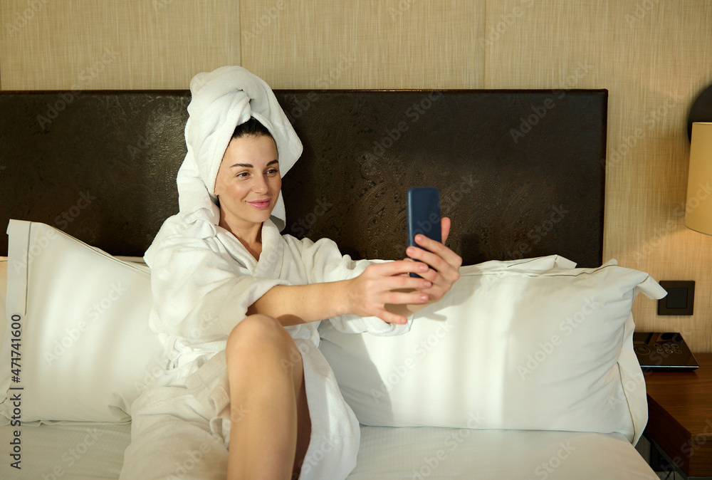 Beautiful dark-haired young woman in white bathrobe and her head wrapped in a towel, relaxing after bathing, lying on a bed in bedroom and smiles cutely taking a selfie Beautiful dark-haired young woman in white bathrobe and her head wrapped in a towel, relaxing after bathing, lying on a bed in bedroom and smiles cutely taking a selfie