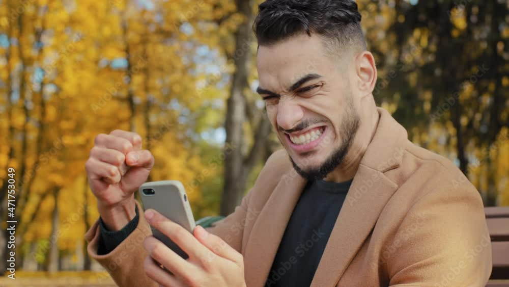 Excited hispanic guy reading good news on smartphone sitting on bench in autumn park young man happily surprised to receive message overjoyed male winner celebrating victory holding telephone outdoors Excited hispanic guy reading good news on smartphone sitting on bench in autumn park young man happily surprised to receive message overjoyed male winner celebrating victory holding telephone outdoors