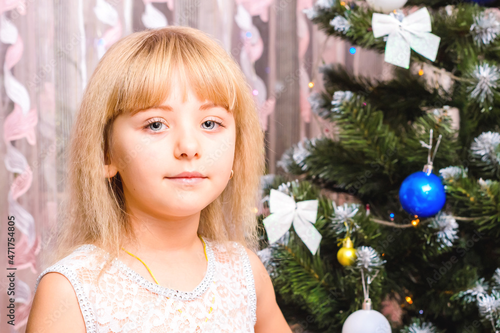 The girl at the Christmas tree in a white dress. A child in a festive mood sits by a decorated Christmas tree.