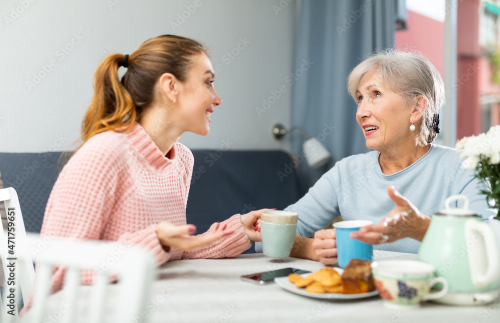 Caucasian young woman and her mature mother sitting at table, talking and drinking tea.