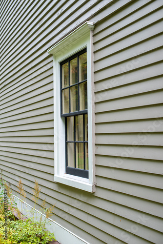 The exterior of a tan color wall of a building. The surface is wood verticle clapboard cape cod siding. There s a vintage multi-pane double hung window in the center with black and white wood trim. 